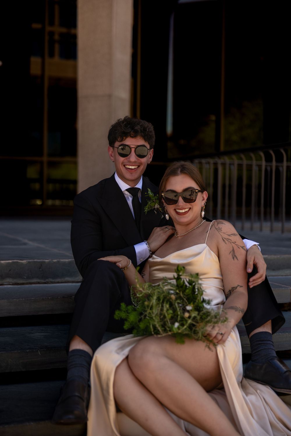 Smiling newlywed couple sitting on steps, bride in gold satin dress holding greenery bouquet, groom in black suit, both wearing sunglasses.