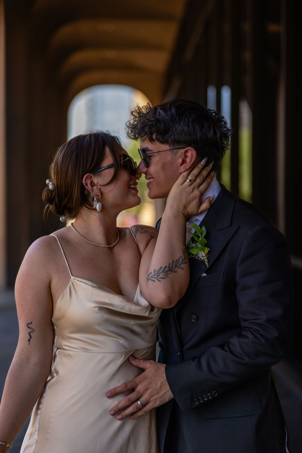 Bride and groom sharing an intimate almost-kiss, she in gold slip dress with tattoos, he in dark suit with floral boutonniere.