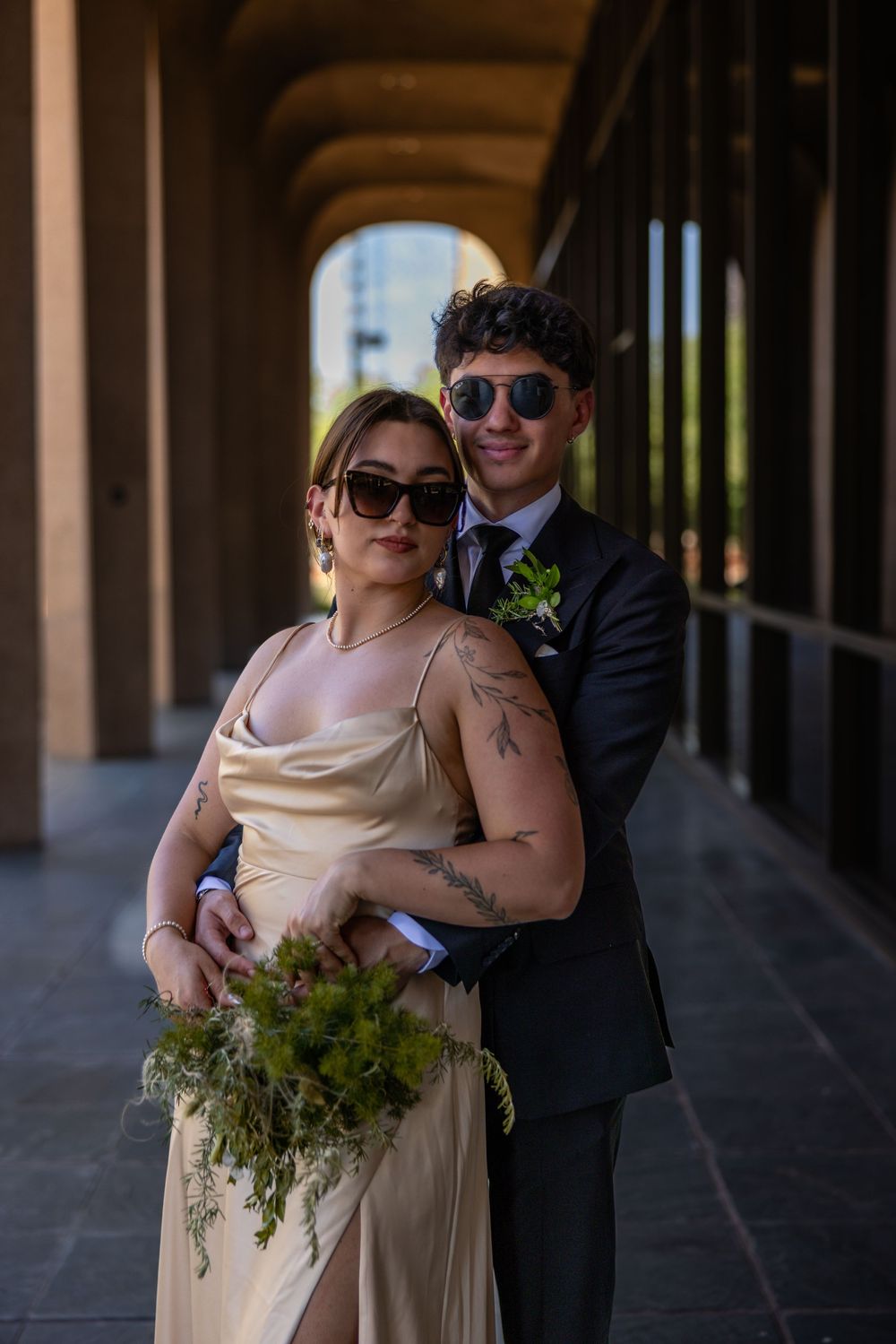 Wedding couple posing in arched corridor, bride in champagne satin dress holding green bouquet, groom in dark suit with sunglasses.