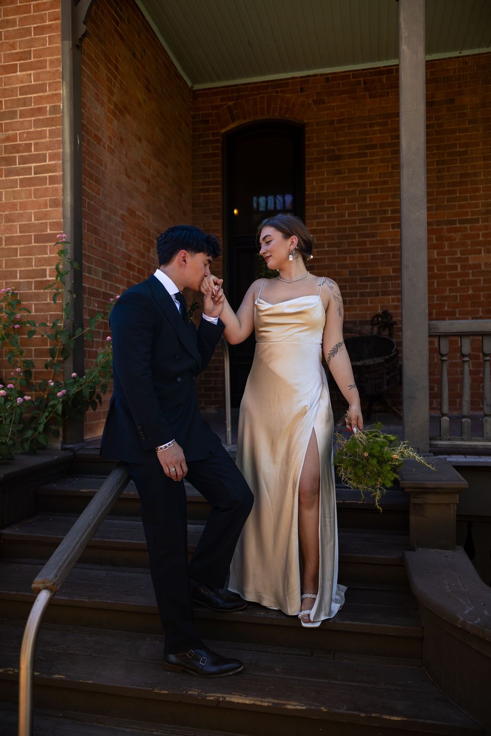 Groom in navy suit kissing bride's hand on historic brick building steps, bride in gold satin gown holding bouquet.
