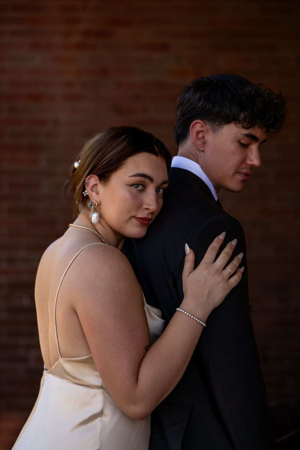 Bride in champagne satin dress with pearl earrings embracing groom from behind against warm brick wall background.
