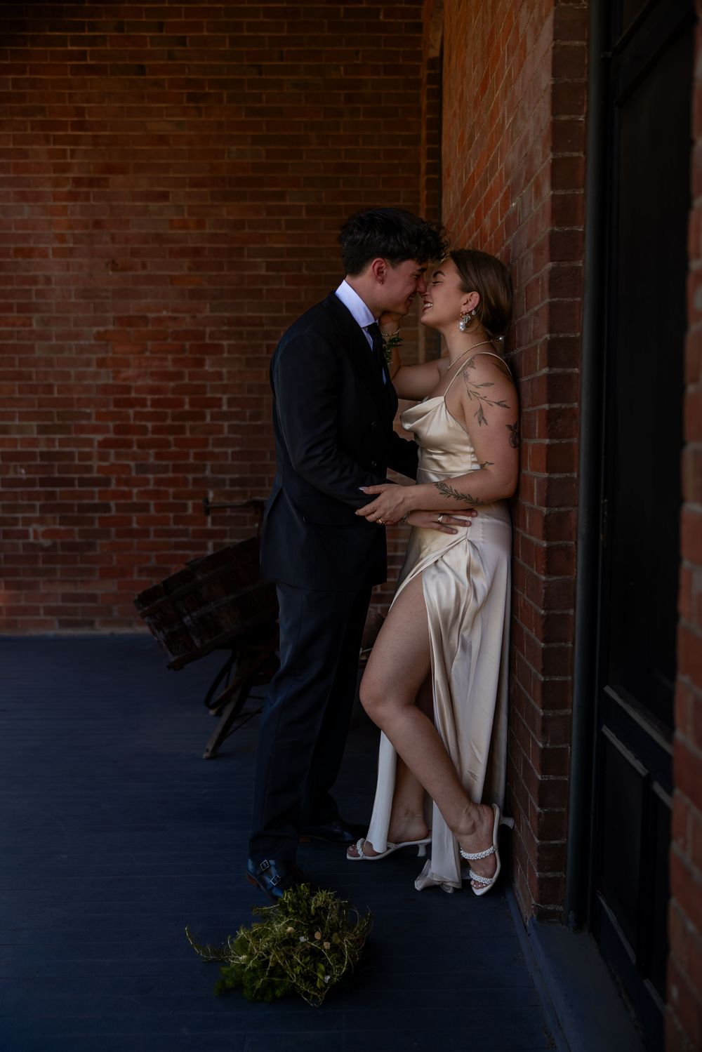 Newlyweds sharing a passionate kiss against rustic brick wall, bride in gold satin slit dress, bouquet on floor.