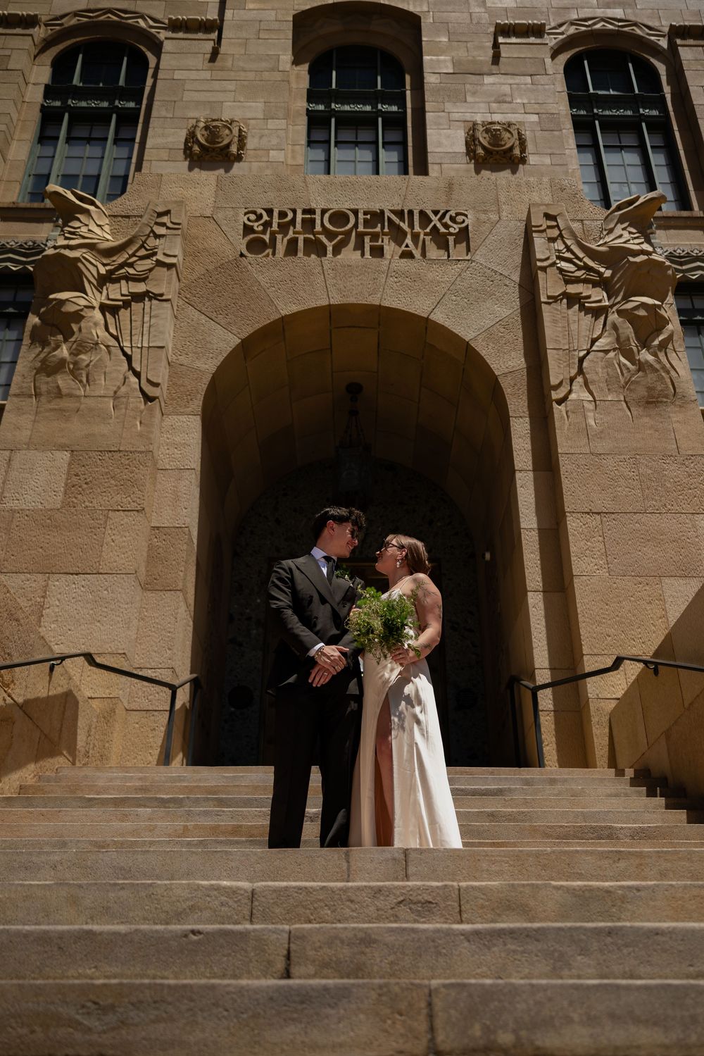 Newlyweds stand together on steps of historic Phoenix City Hall holding green bouquet.