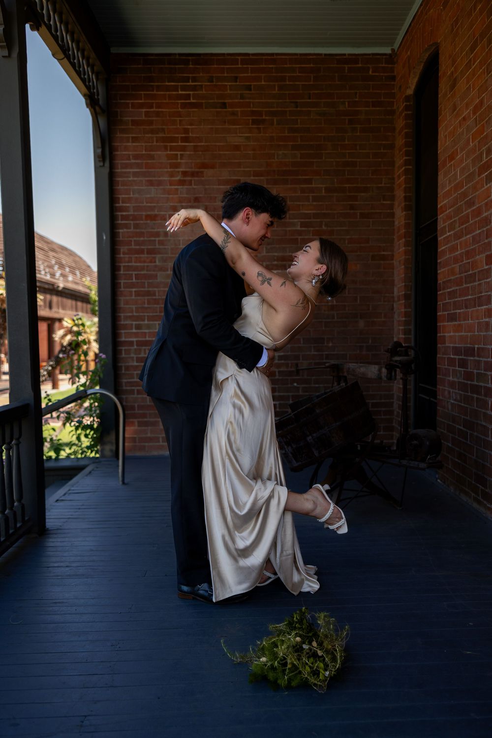 Groom lifting bride in gold satin dress on blue wooden porch of brick building, bouquet dropped on floor.