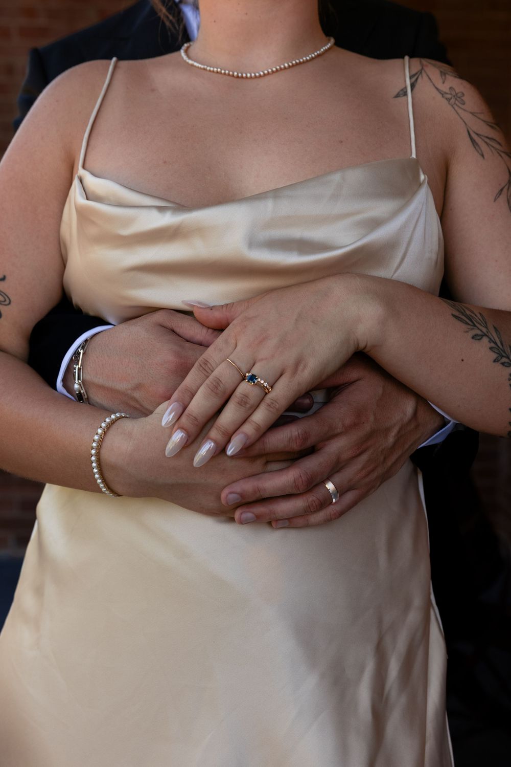 Close-up of bride and groom's hands clasped together showing wedding rings, bride in gold satin dress with pearl bracelet.