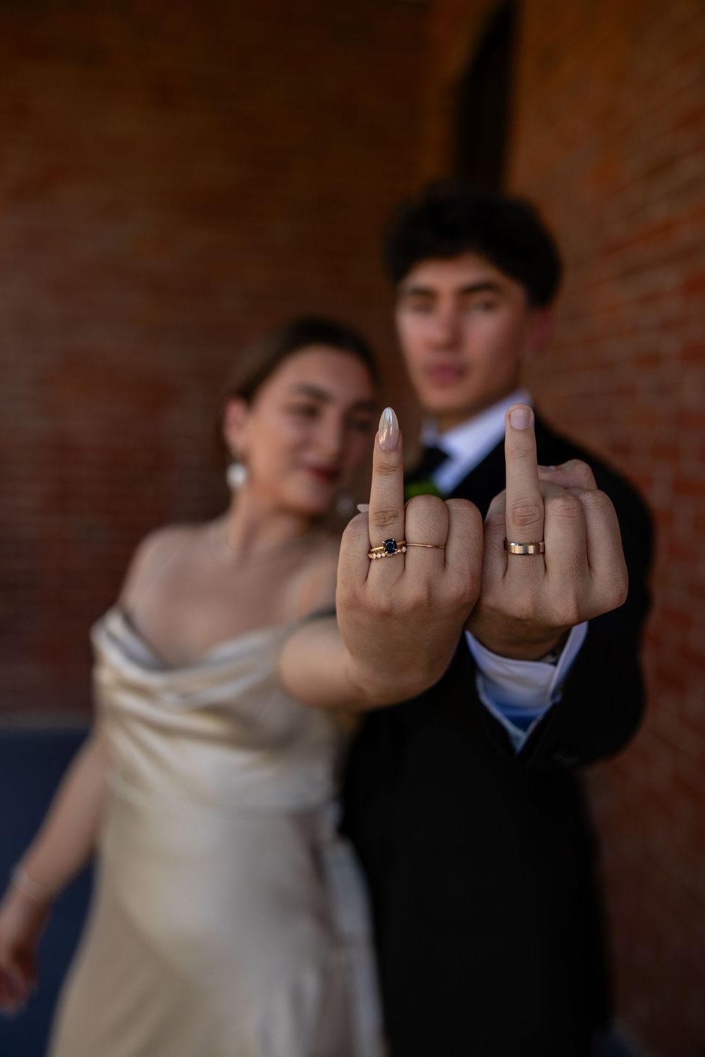 Bride and groom showing off wedding rings on raised middle fingers, brick wall background, playful wedding photo.