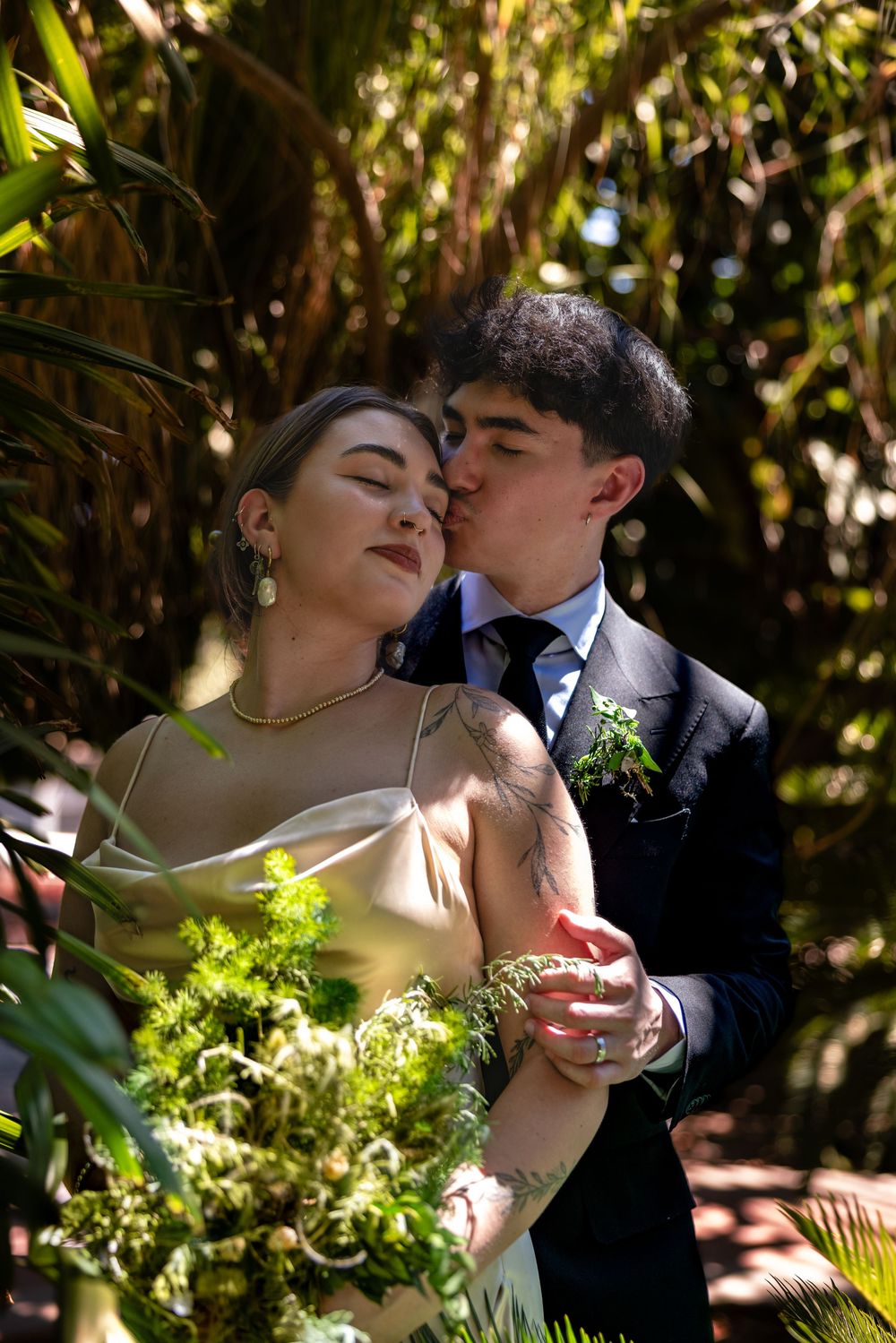 Groom kissing bride's temple in lush garden setting, bride holding green botanical bouquet in champagne slip dress.