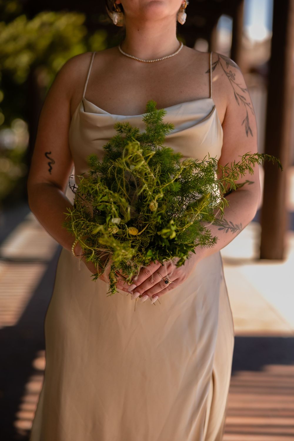Tattooed bride in champagne slip dress holding lush green botanical bouquet at outdoor wedding ceremony.
