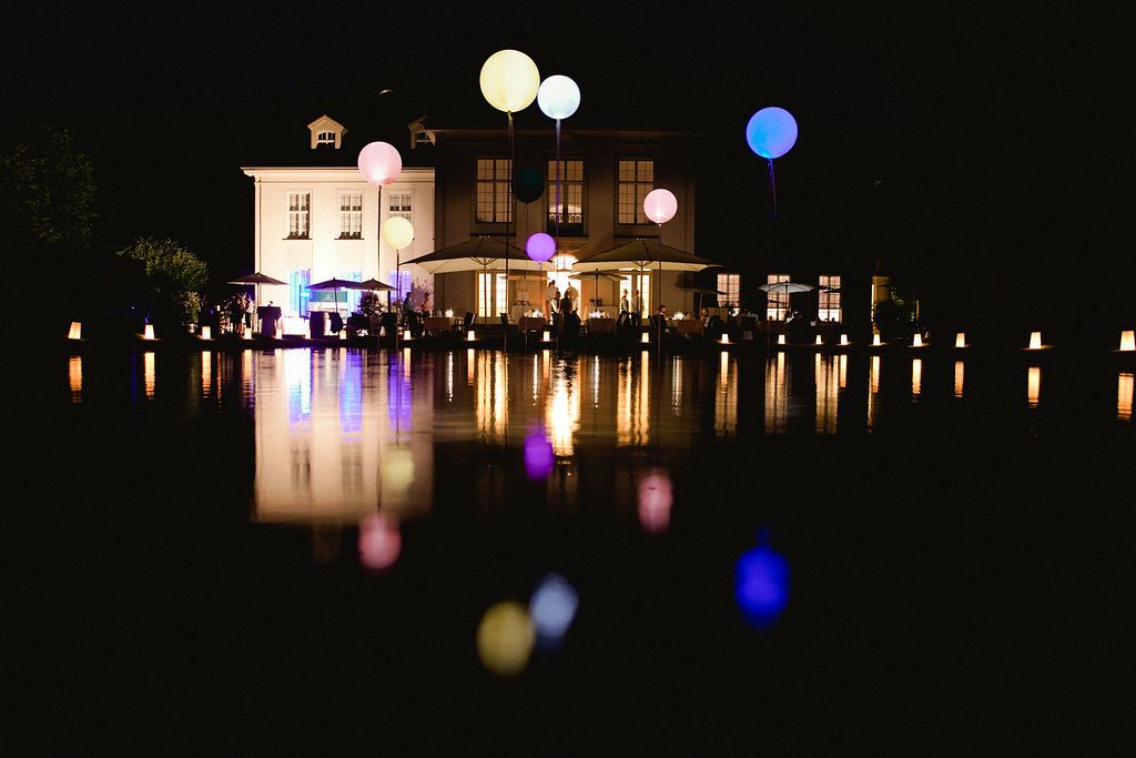 Illuminated building with glowing balloons reflecting in calm water at night, creating a magical mirrored effect.