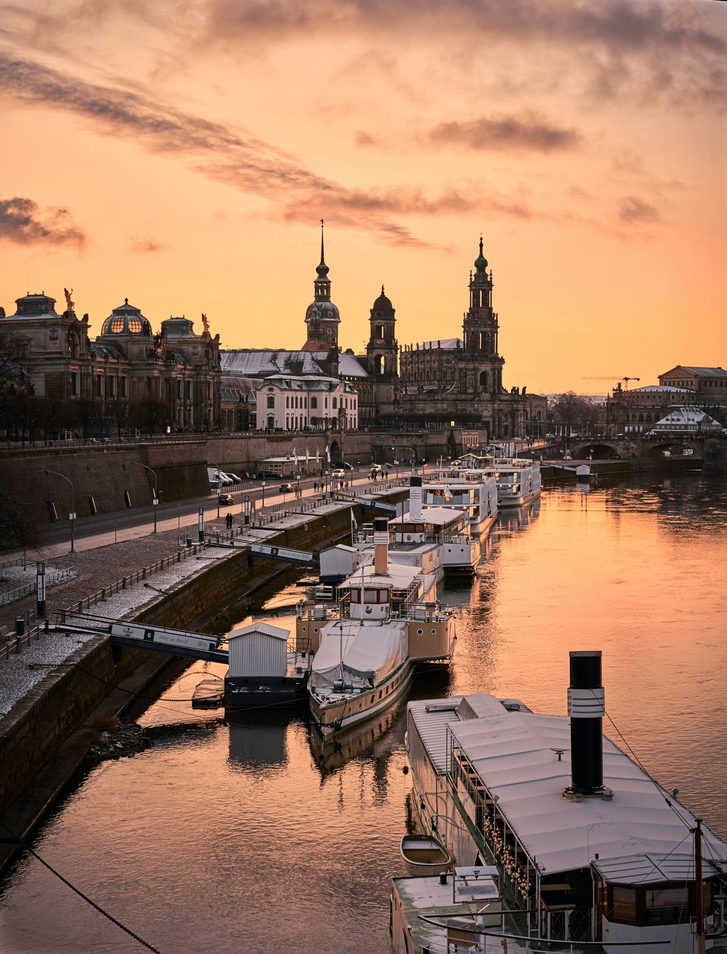 Historic Dresden skyline with boats docked along the Elbe River during a golden sunset.