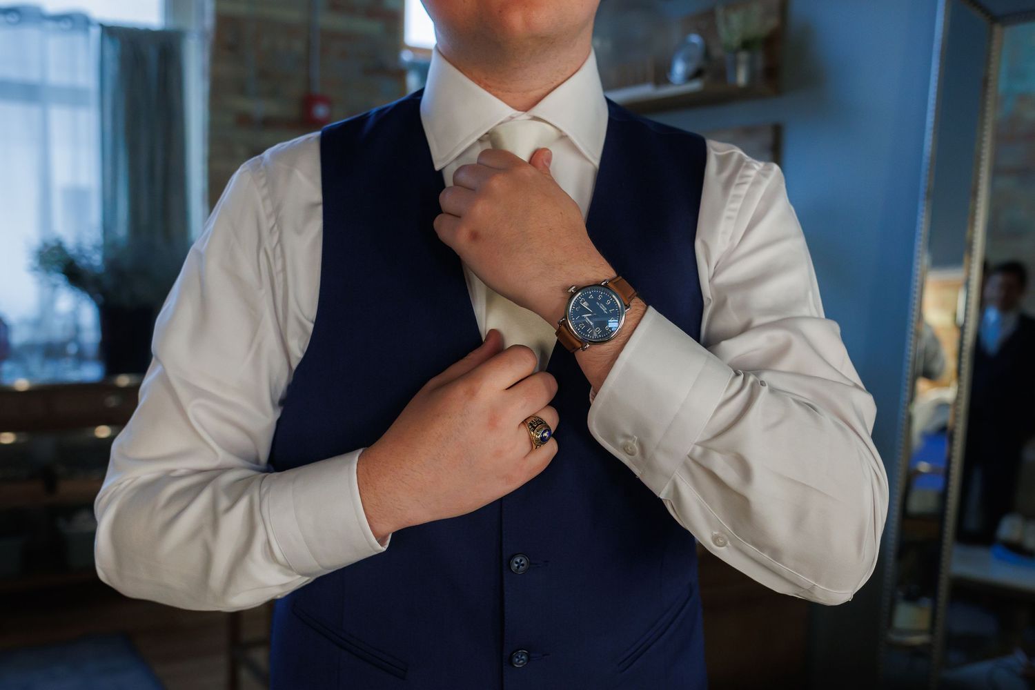 Professional adjusting a blue necktie while wearing a white dress shirt and navy vest in an office setting.