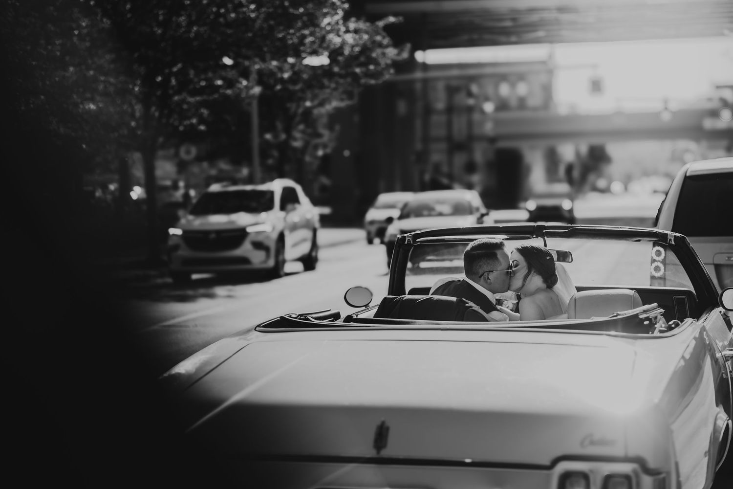 A classic white convertible car drives down a city street in black and white photography.