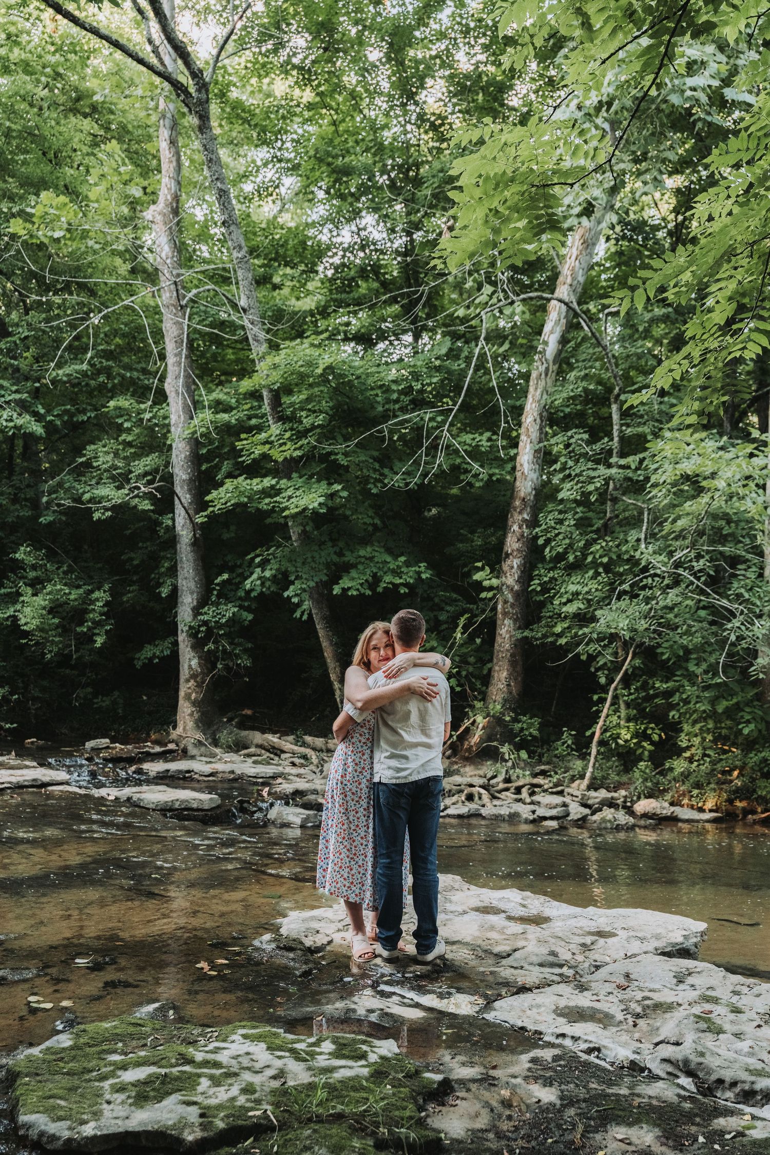 A couple stands close together sharing an intimate moment by a scenic creek in a lush green forest setting.