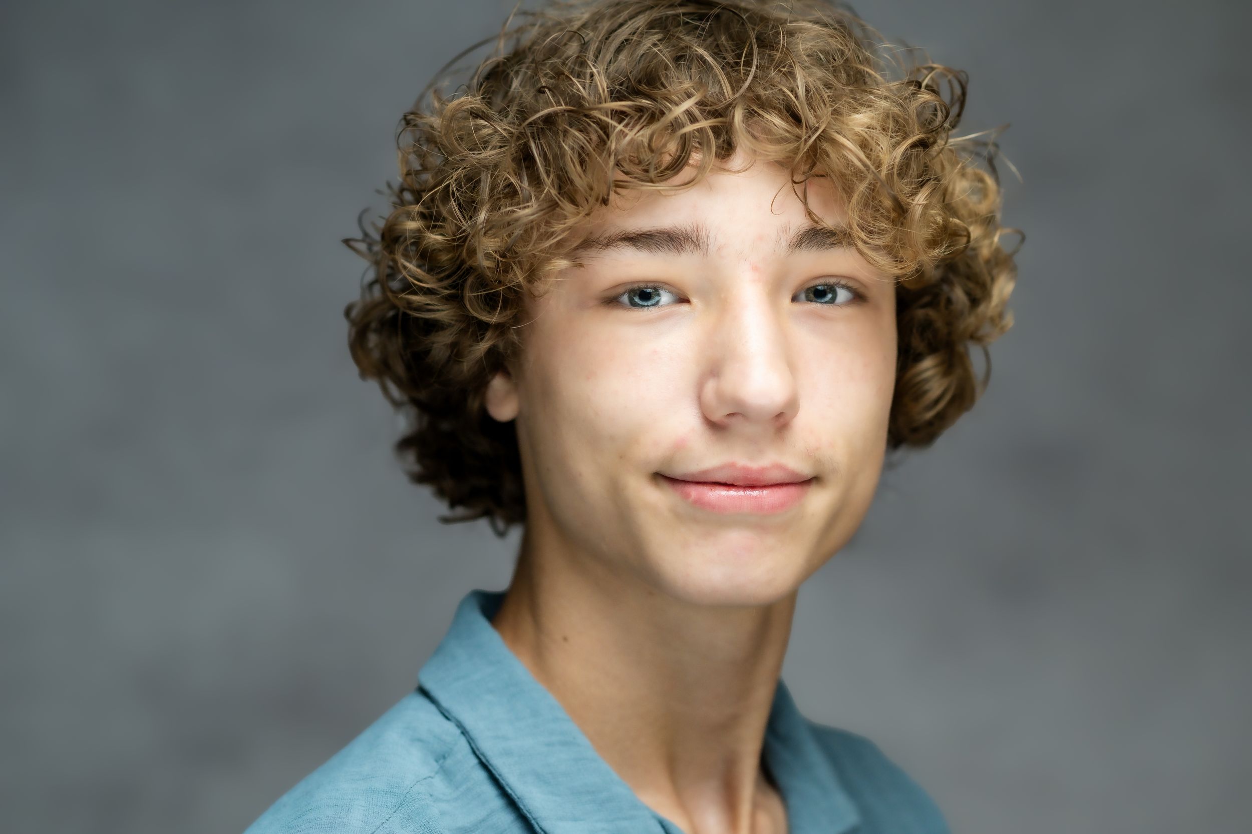 Someone with curly hair wearing a light blue shirt smiles warmly against a gray studio background.