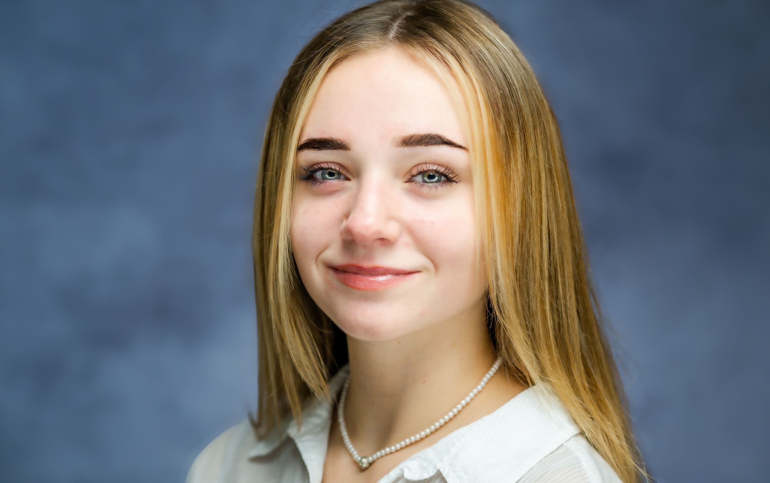 Professional headshot portrait of a young person with long blonde hair wearing a white button-up shirt against a blue backdrop.