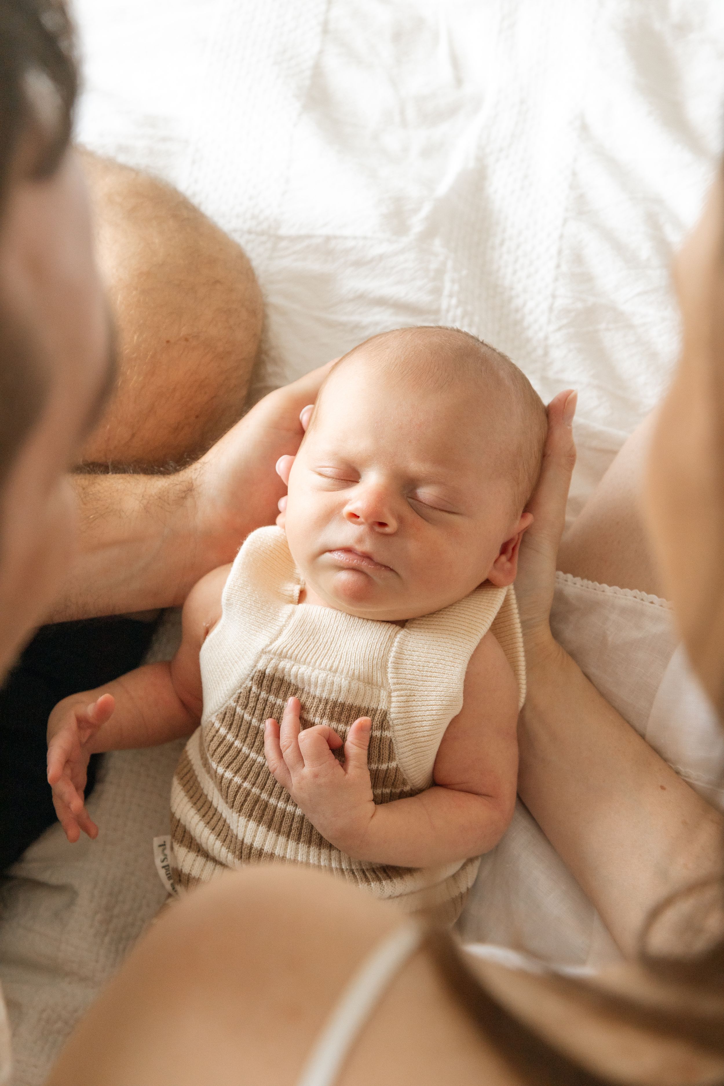 A sleeping infant wearing a cream-colored romper lies peacefully on white bedding while hands reach towards them.