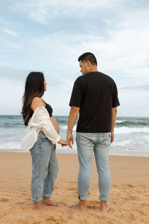 A couple holding hands while standing barefoot on a sandy beach with ocean waves in the background.