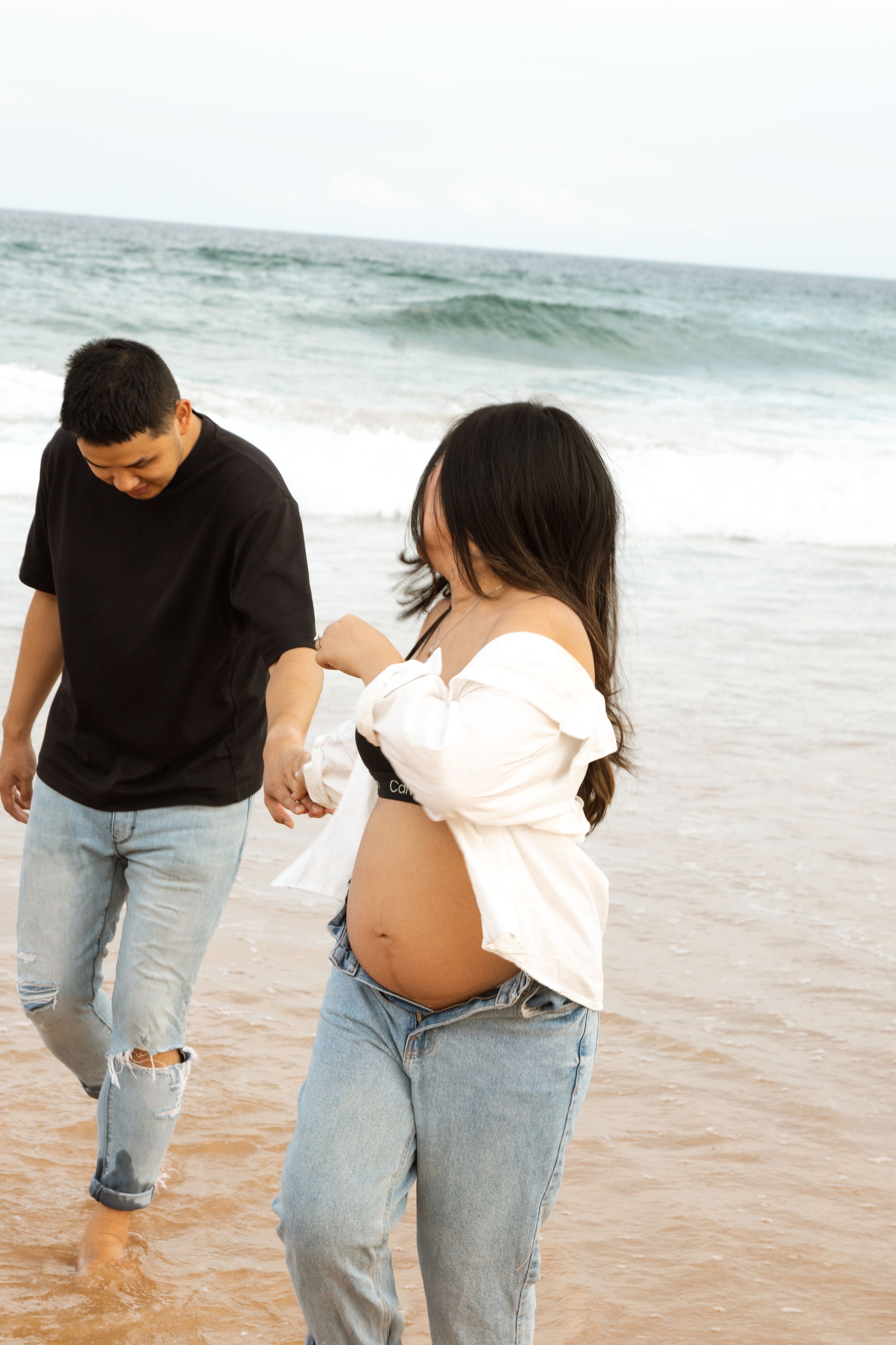 Couple walking on beach holding hands with pregnant woman in white top and jeans along the ocean shore.