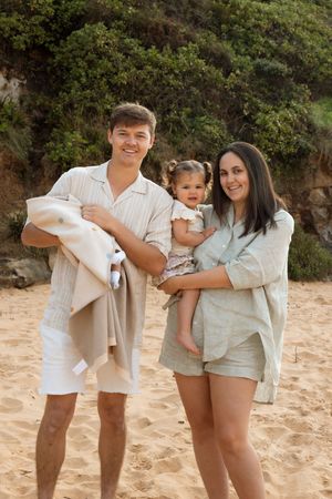 Family wearing coordinated beige and white outfits poses together on sandy beach with greenery in background.