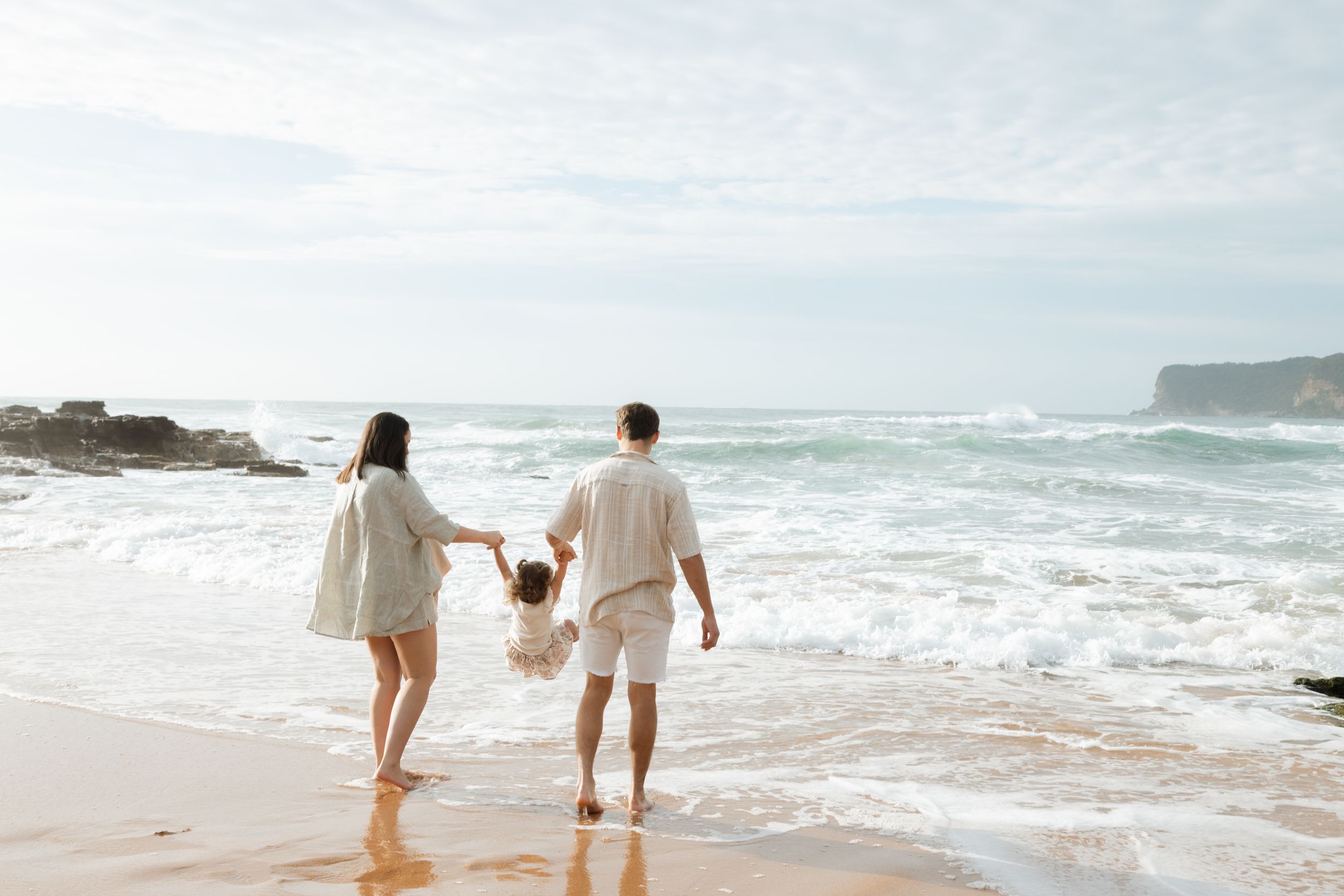 A couple in white outfits walking hand in hand on a scenic sandy beach with ocean waves in the background.