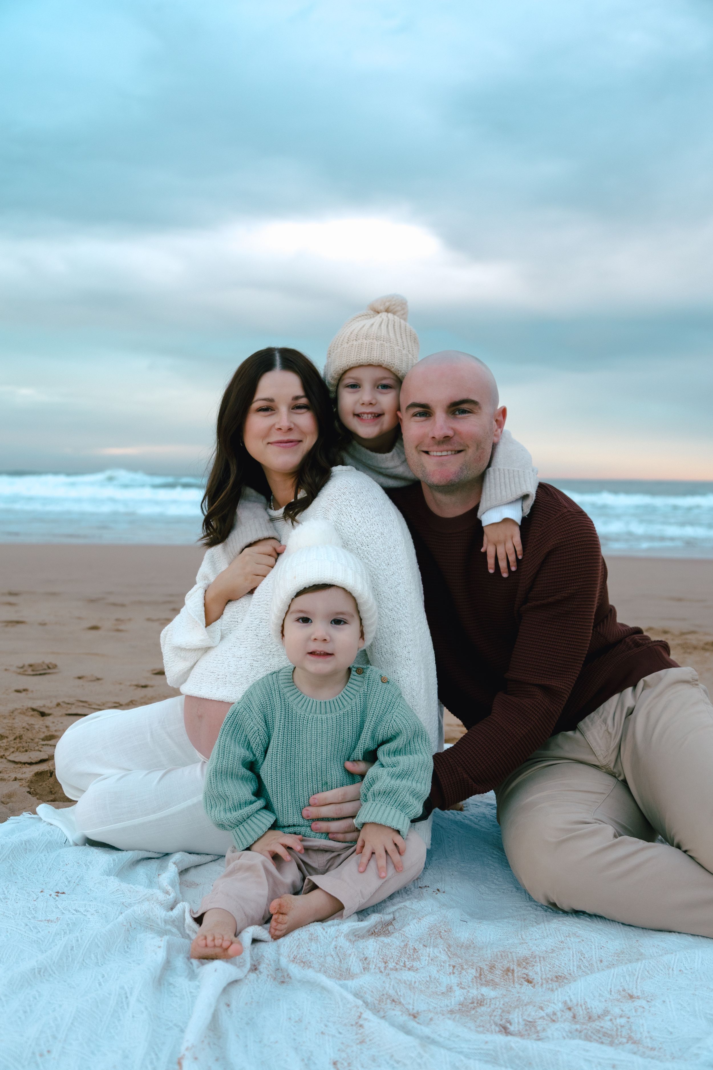 A family relaxes together on a blanket at the beach during sunset with soft cloudy skies and ocean waves in background.