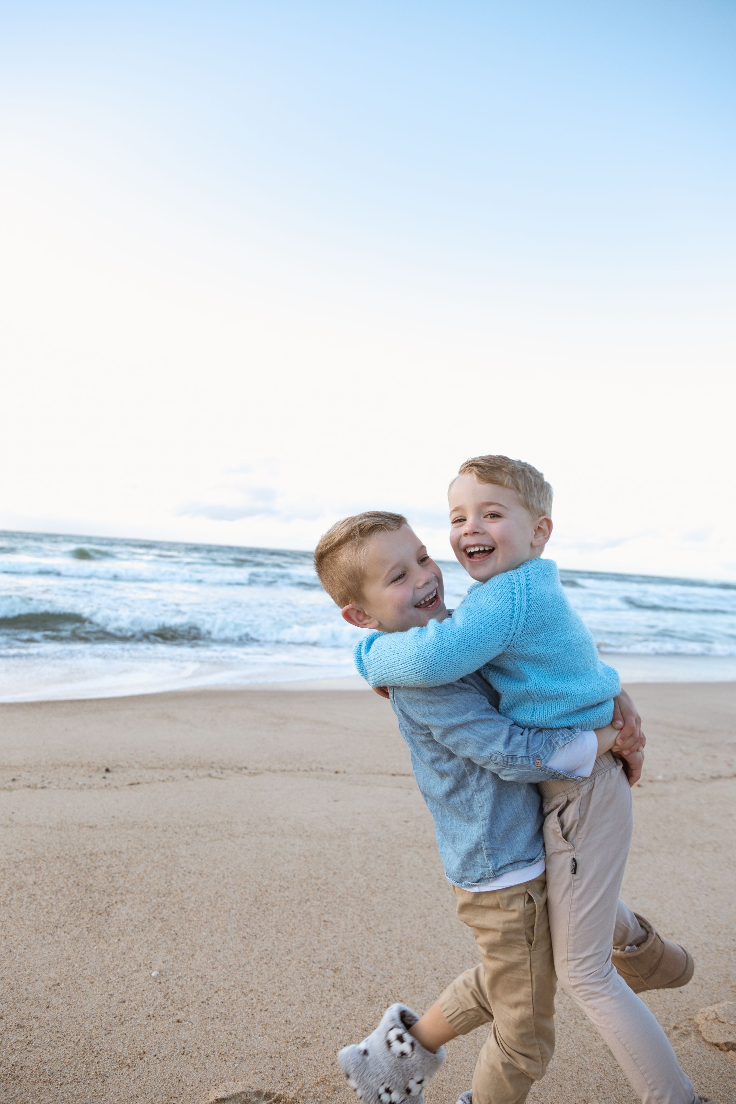 Two children in blue sweaters play and embrace joyfully on a sandy beach at sunset with ocean waves in background.