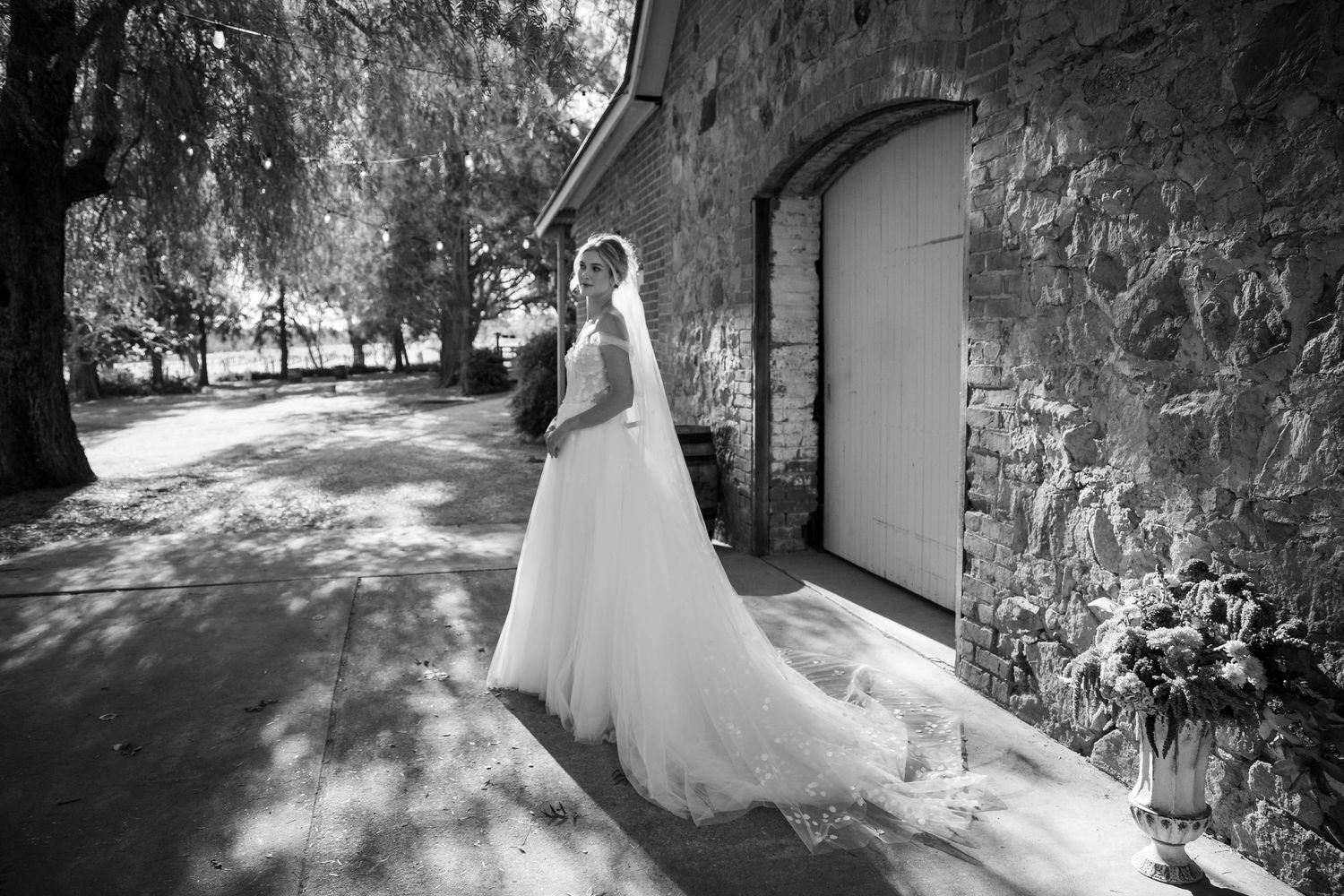 A bride in a flowing white dress stands in dappled sunlight next to a stone wall with an arched doorway.