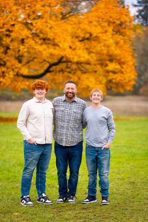 Three people stand in a line on green grass with vibrant orange autumn foliage in the background.