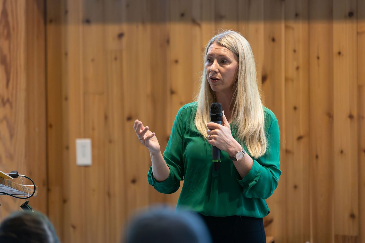 A presenter in a bright green sweater speaks while gesturing with both hands in front of a wooden paneled wall.