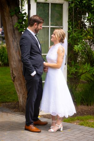 A bride in a tea-length lace dress and groom in dark suit share an intimate moment in a garden setting on their wedding day.