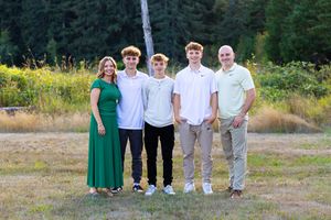 A group of five people standing together outdoors in a grassy field with evergreen trees in the background.