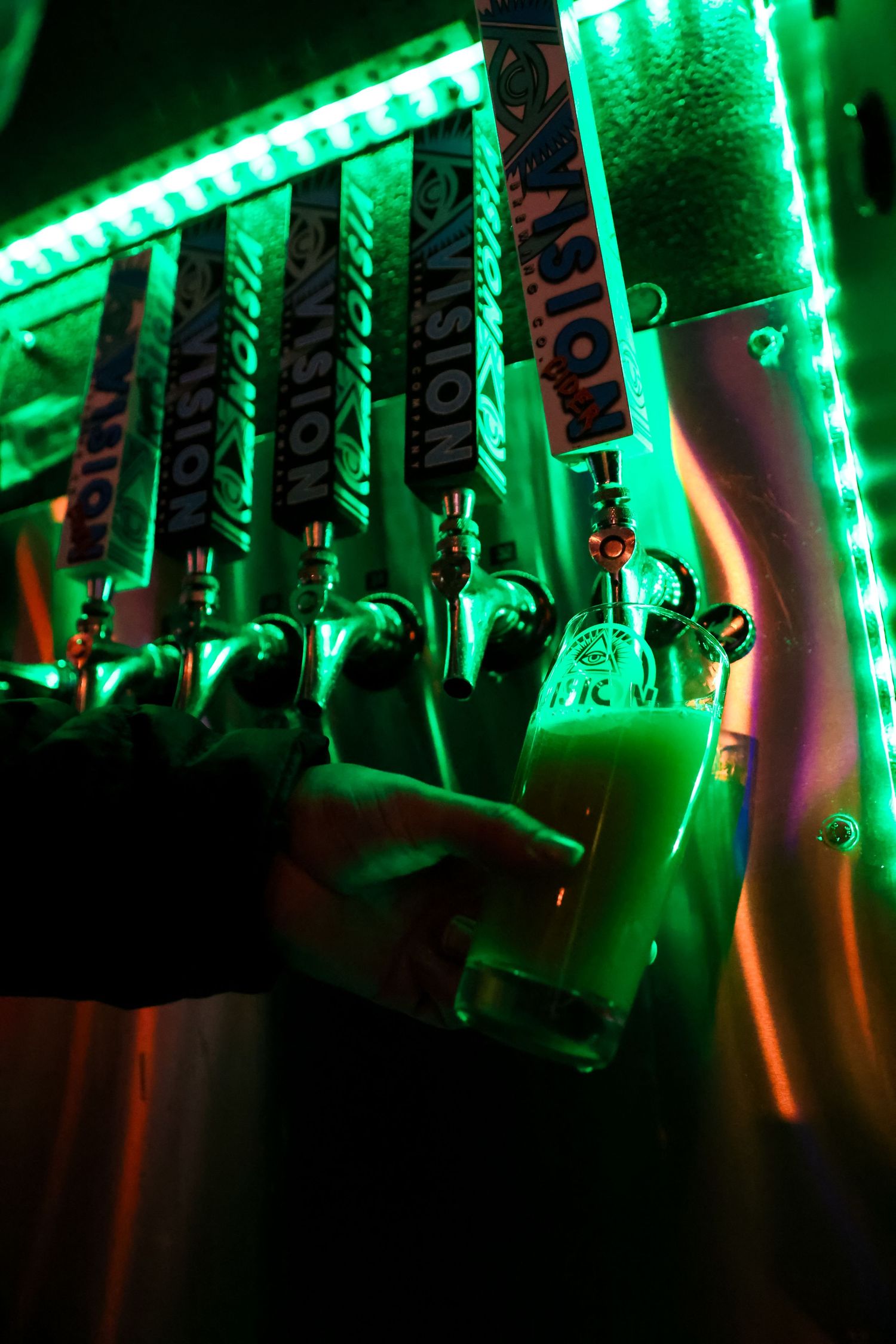 Beer tap handles illuminated by vibrant green LED lighting at a bar with stainless steel fixtures.