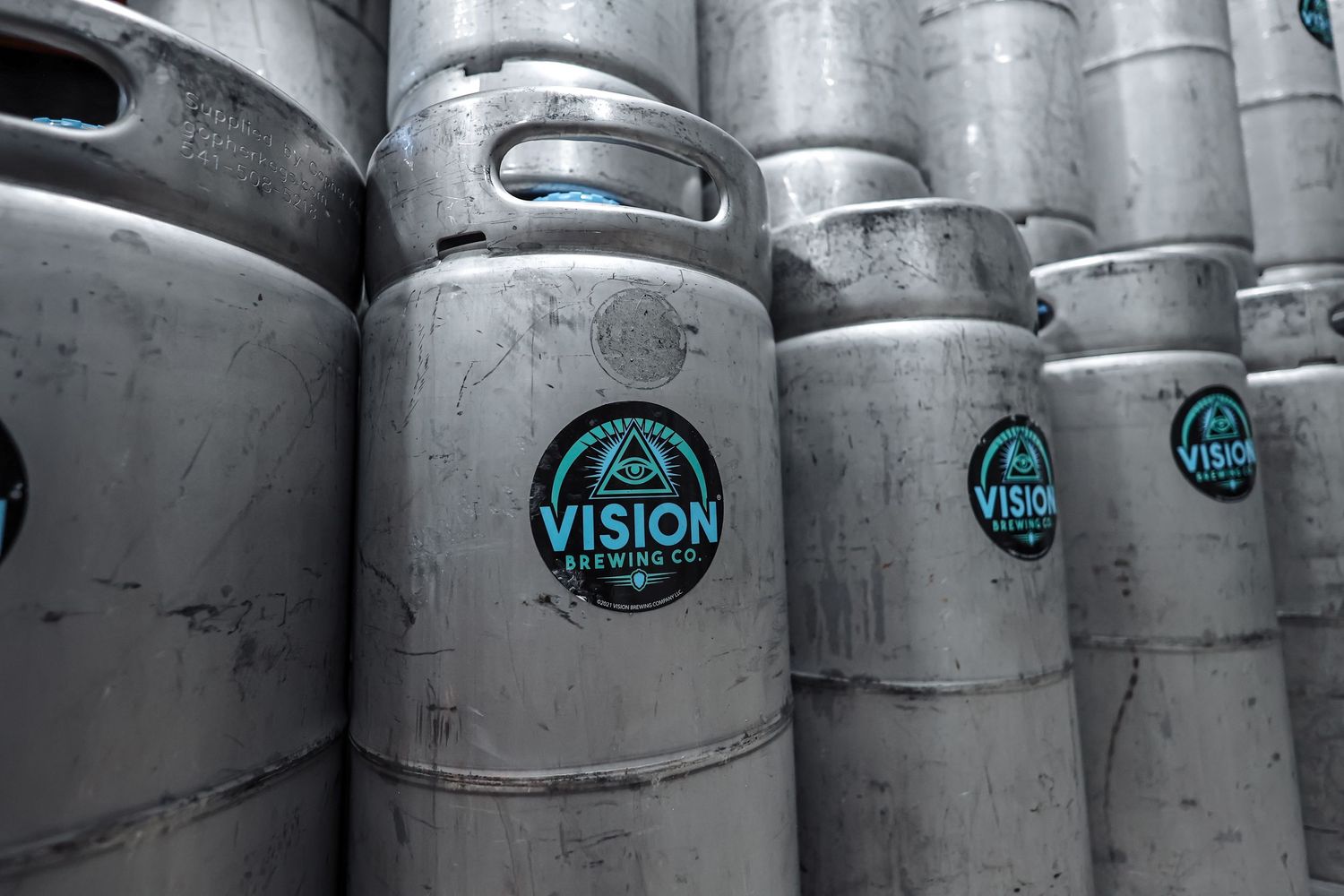 Row of stainless steel beer kegs with Vision Brewing Company logo stacked in brewery storage area.