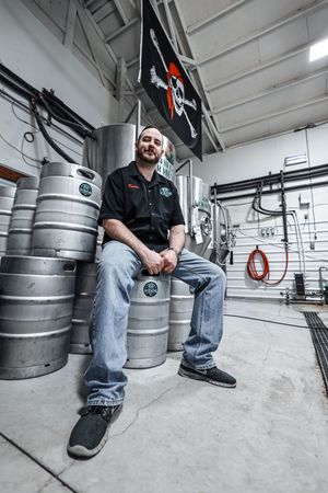 Brewery worker sits on stainless steel kegs in industrial storage room with pirate flag on wall.