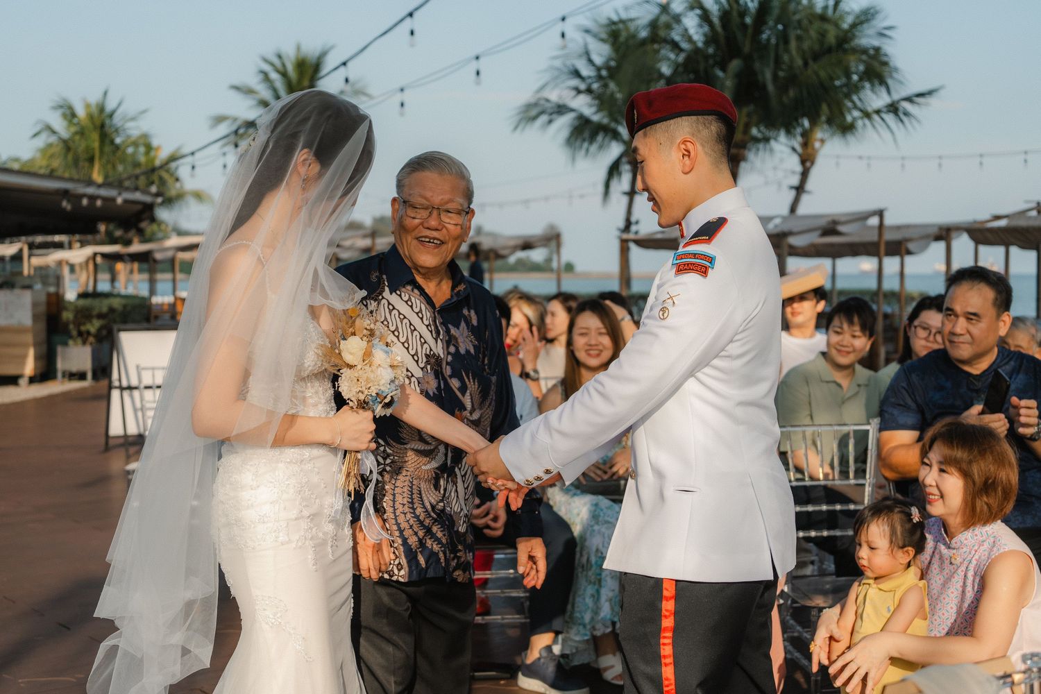 Outdoor wedding ceremony taking place under palm trees with guests watching.