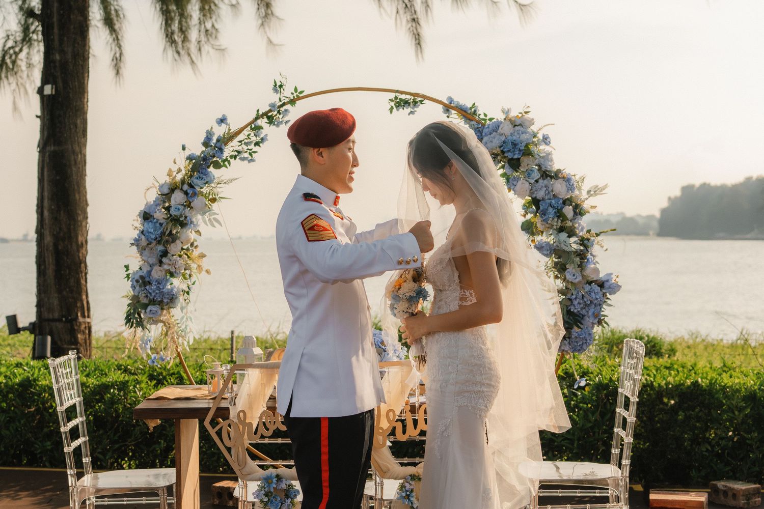 A romantic outdoor wedding ceremony with a floral archway overlooking a waterfront setting at sunset.