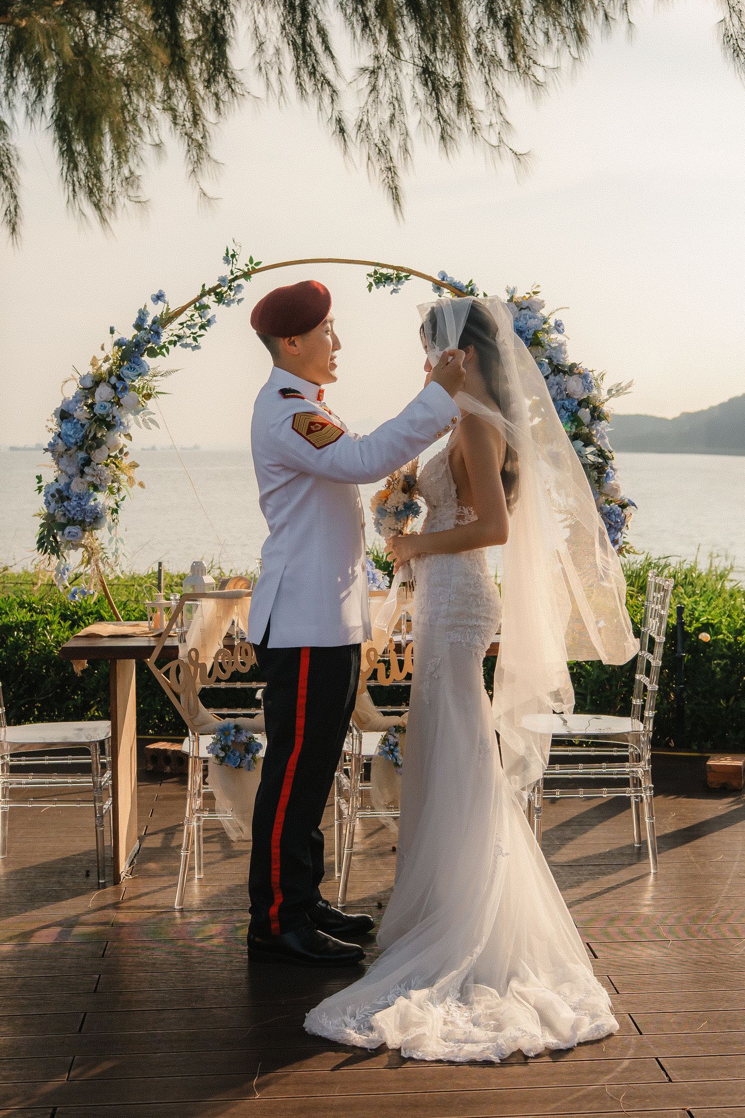 Outdoor beach wedding ceremony with blue floral arch and ocean backdrop at sunset.
