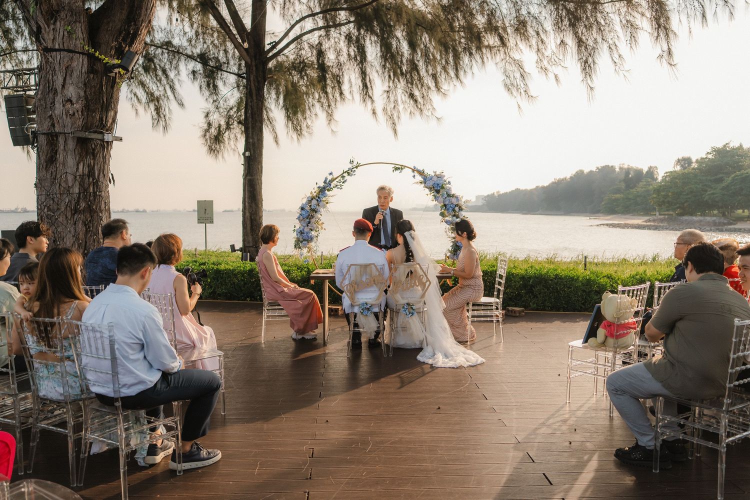 An intimate waterfront wedding ceremony with guests seated around a floral arch on a wooden deck.