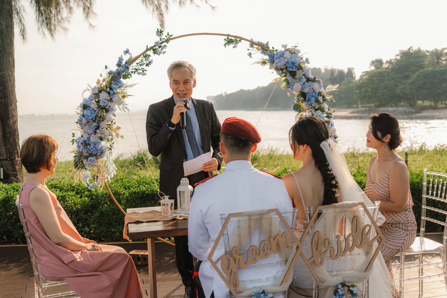 A small wedding party gathered for an outdoor ceremony under a blue floral arch with water views.