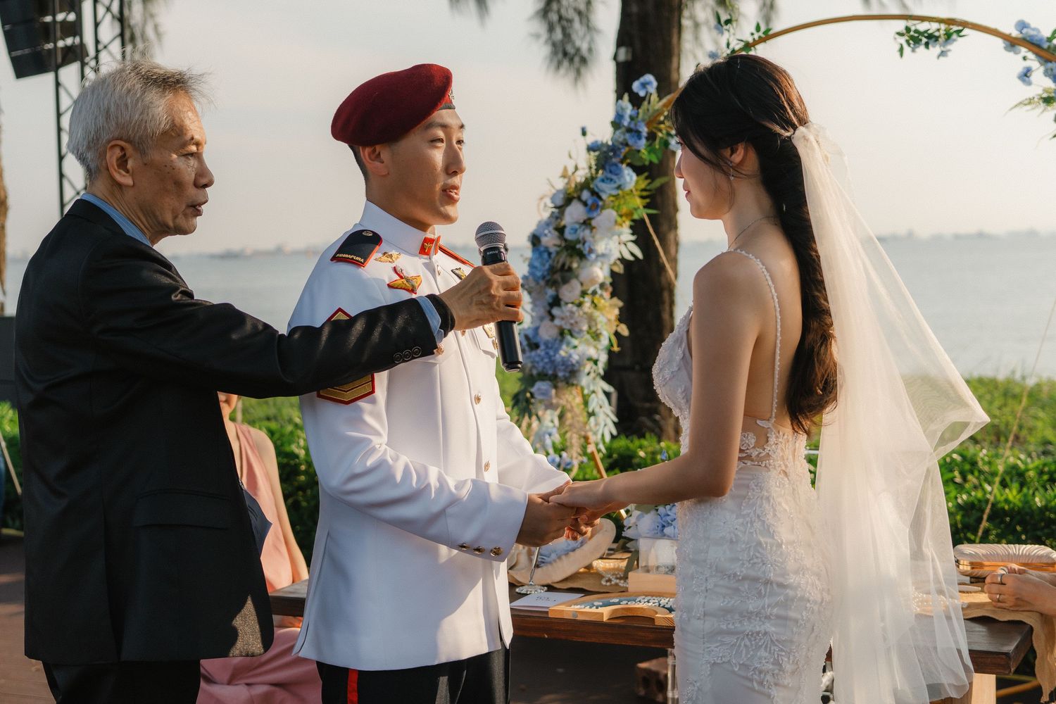 An outdoor wedding ceremony takes place under a floral arch with water views in the background.