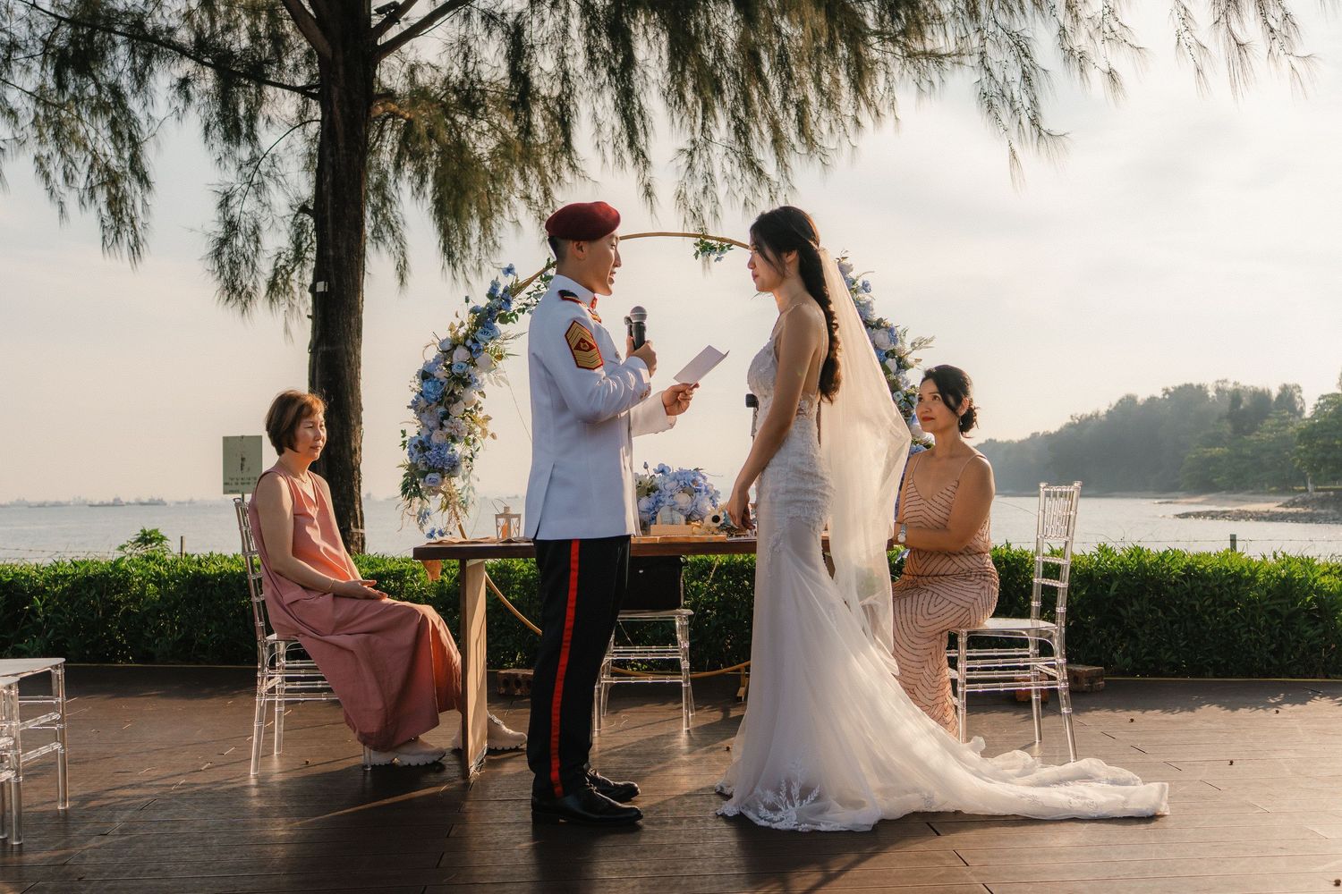 An intimate waterfront wedding ceremony with a small group gathered around a floral arch at dusk.