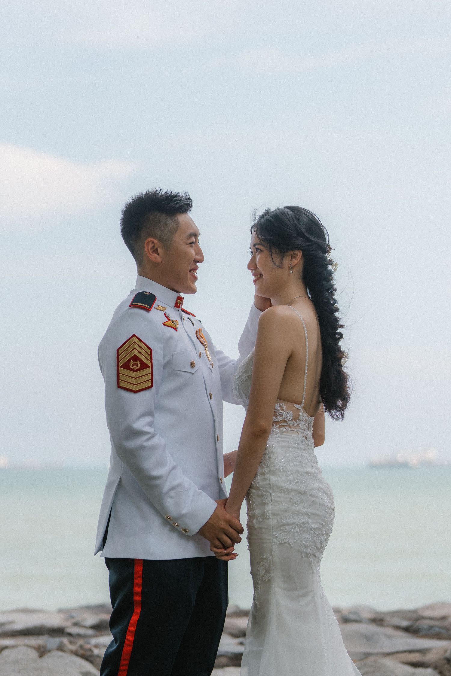 A military serviceman in dress uniform and bride in white lace gown pose together on a rocky beach shore at sunset.