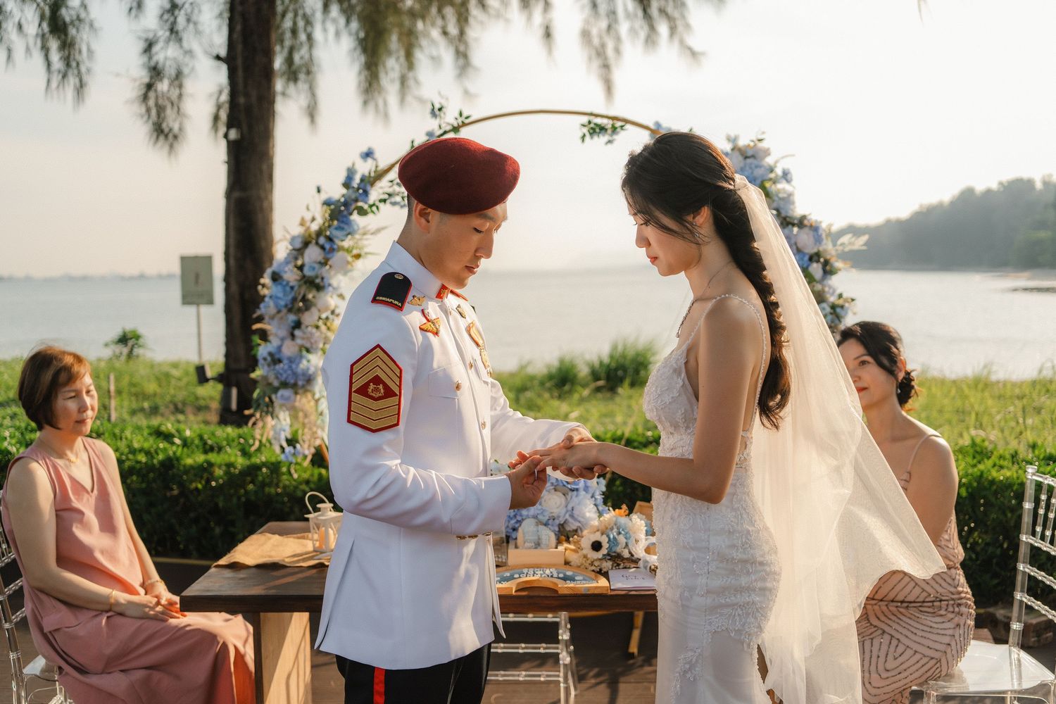 Outdoor waterfront wedding ceremony with floral arch and guests at sunset.