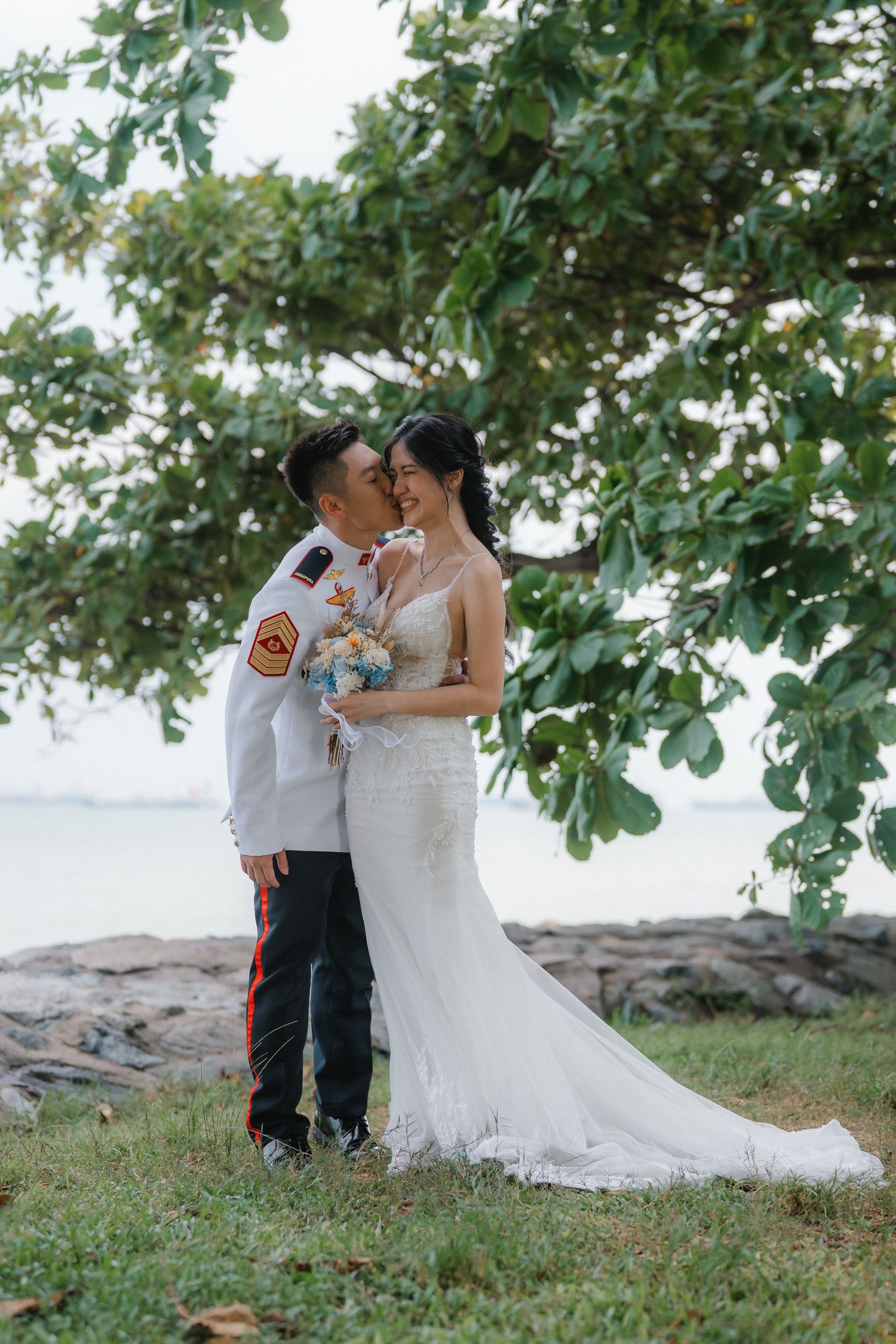 A romantic wedding photo of a couple embracing outdoors with a white lace gown and military uniform under green trees.