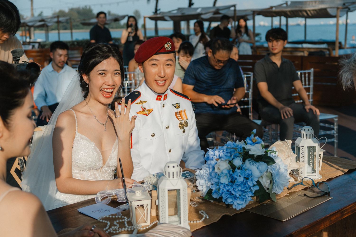 Wedding reception celebration at seaside venue with lantern centerpieces and blue floral arrangements on wooden tables.