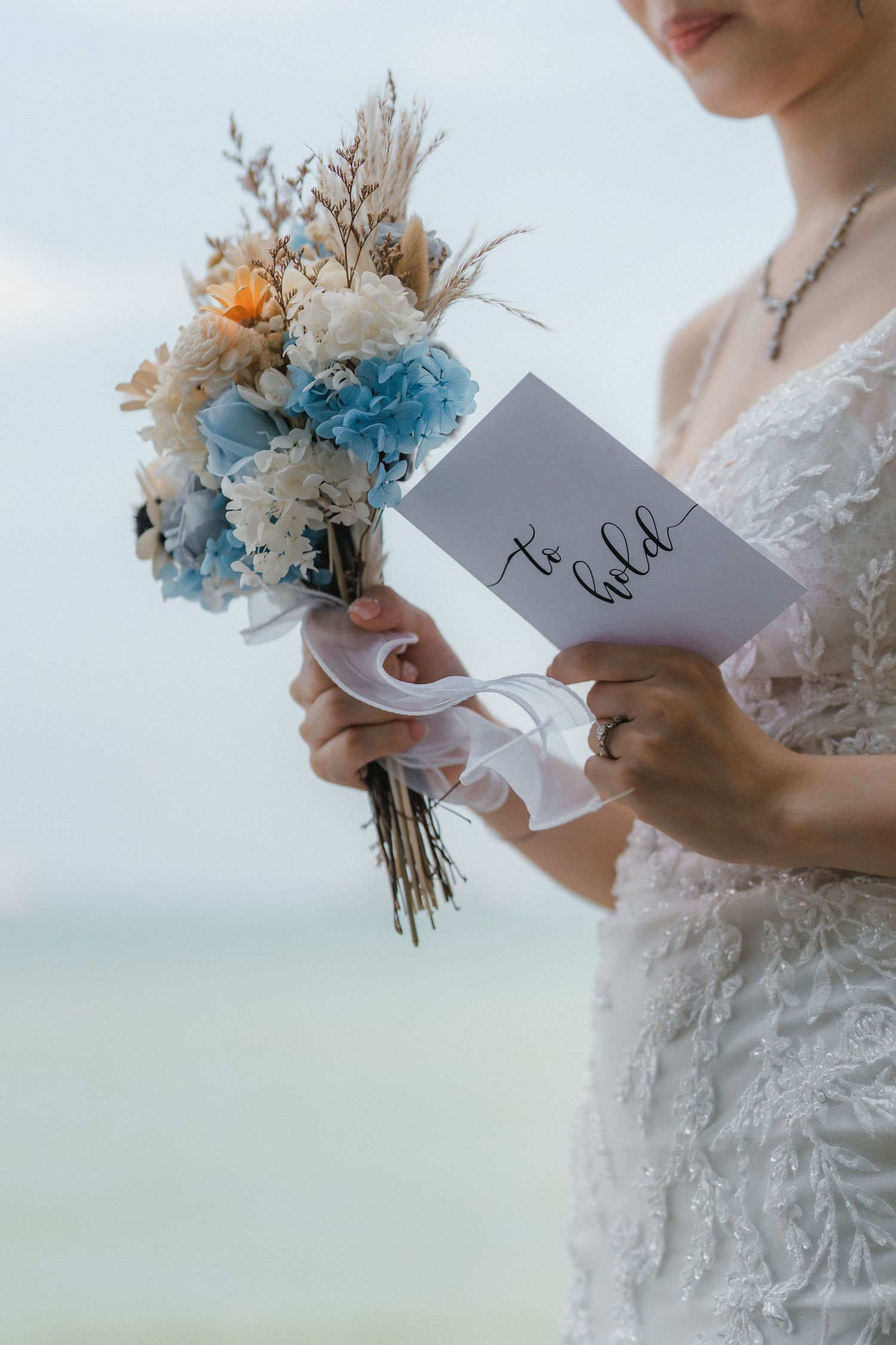 Close up of wedding bouquet with blue and white flowers and handwritten vows card held by someone in lace dress.