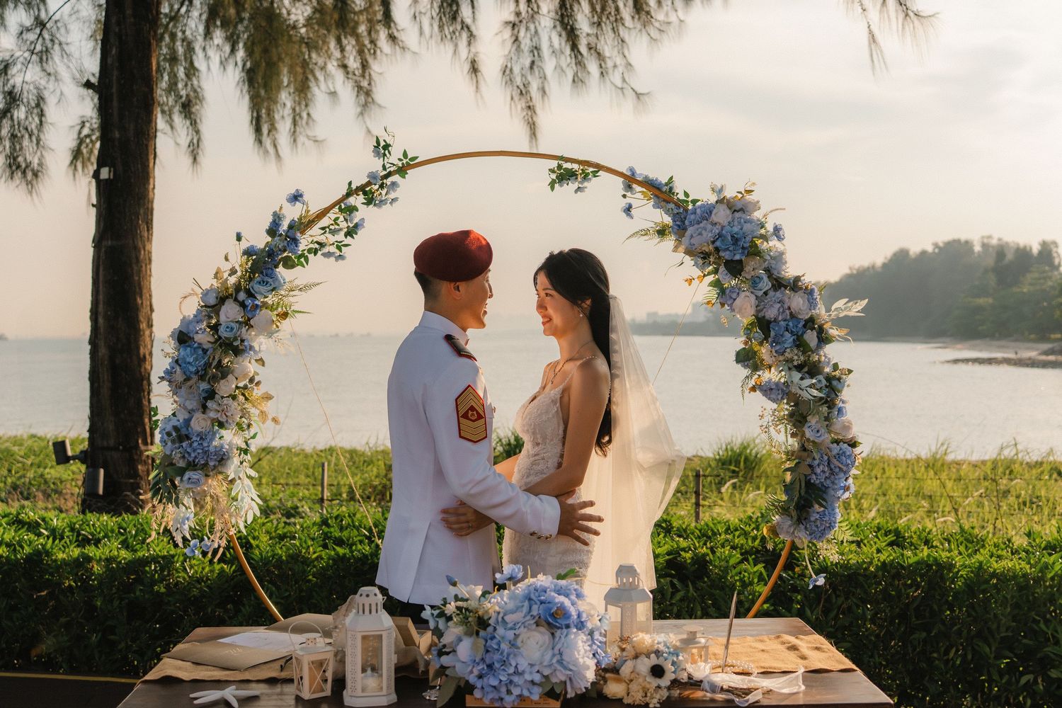 A wedding couple stands beneath a floral arch with blue flowers overlooking the water at sunset.