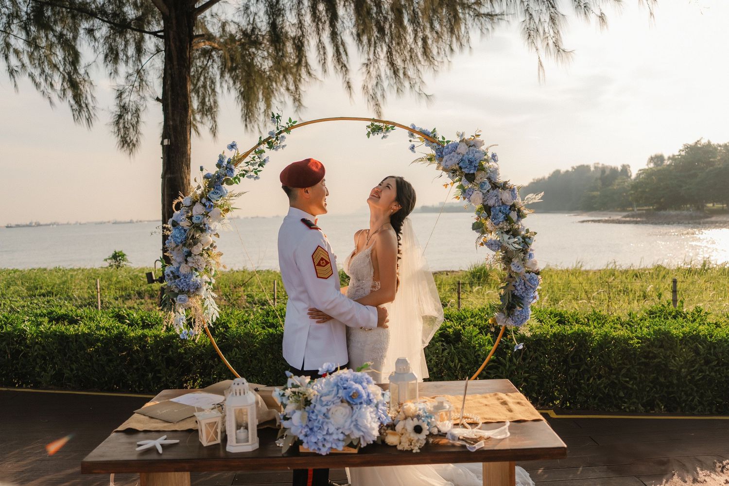 Wedding couple under a floral arch decorated with blue flowers against a waterfront sunset backdrop.