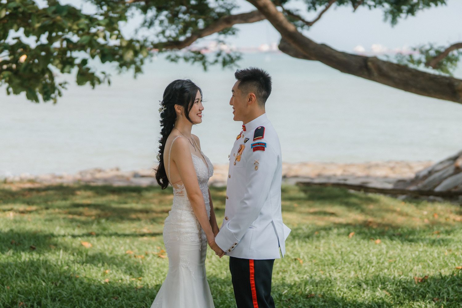 A couple in wedding attire shares an intimate moment under a tree branch at a scenic outdoor venue.