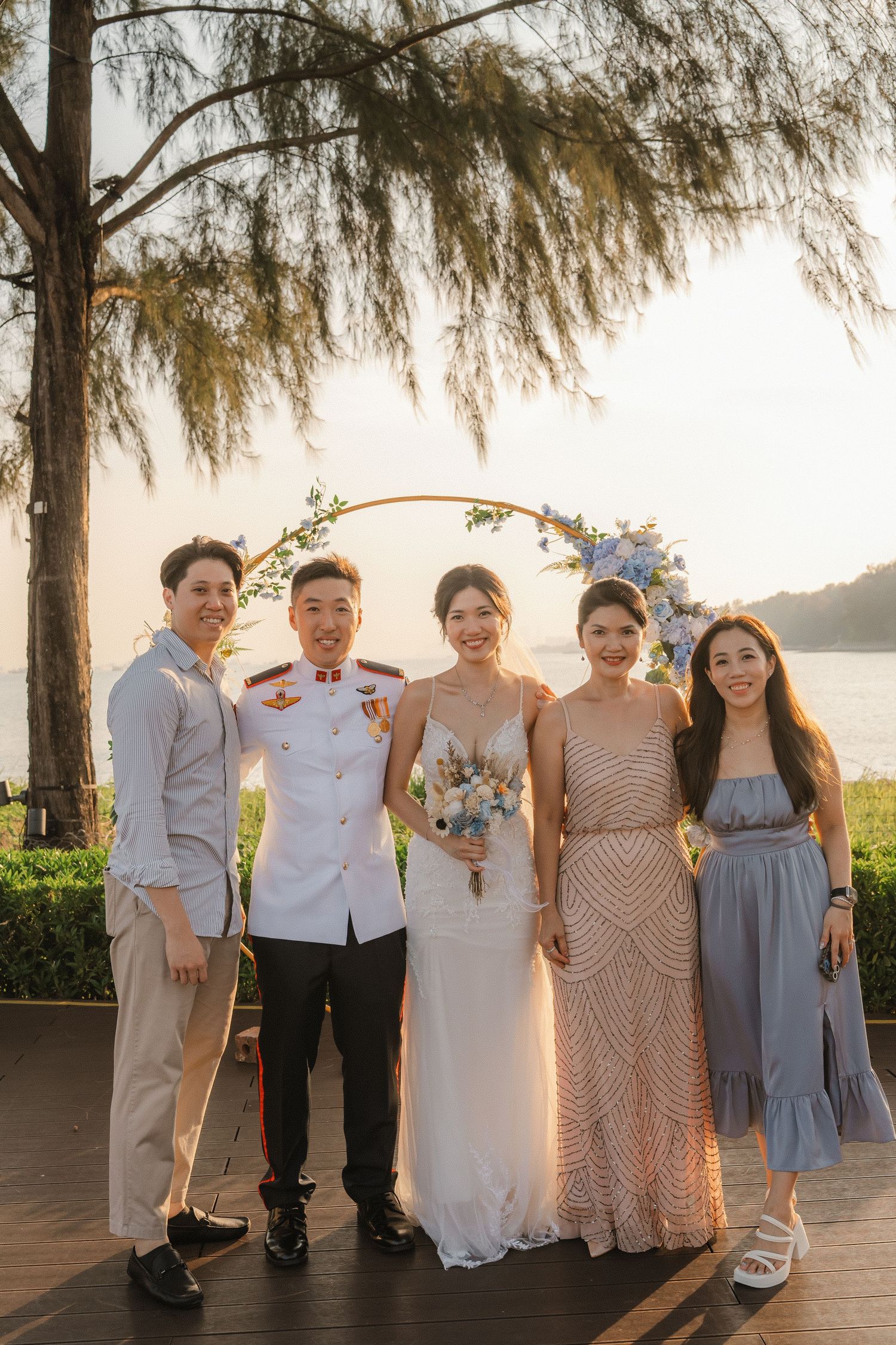 Wedding party poses together at sunset beneath a willow tree on a waterfront deck.